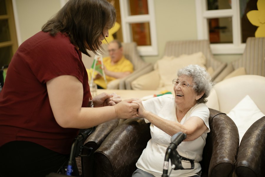 A carer holding hands with a smiling older woman who uses a walking frame in her armchair.