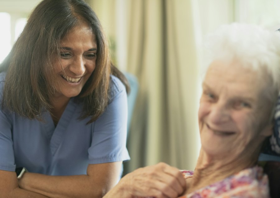 A nurse in a blue tunic sharing a warm conversation with an older woman.