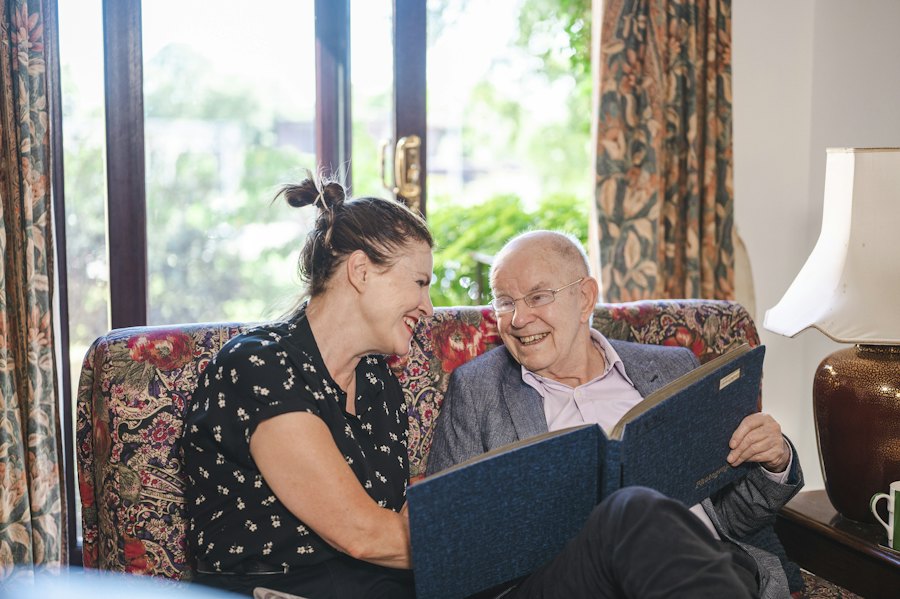 A woman and an older man laughing together over a photo album on a sofa at home.