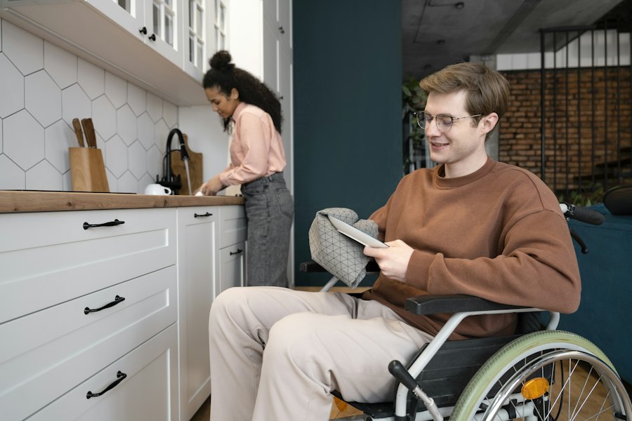 A young man in a wheelchair at home in his kitchen while a support worker prepares food nearby.