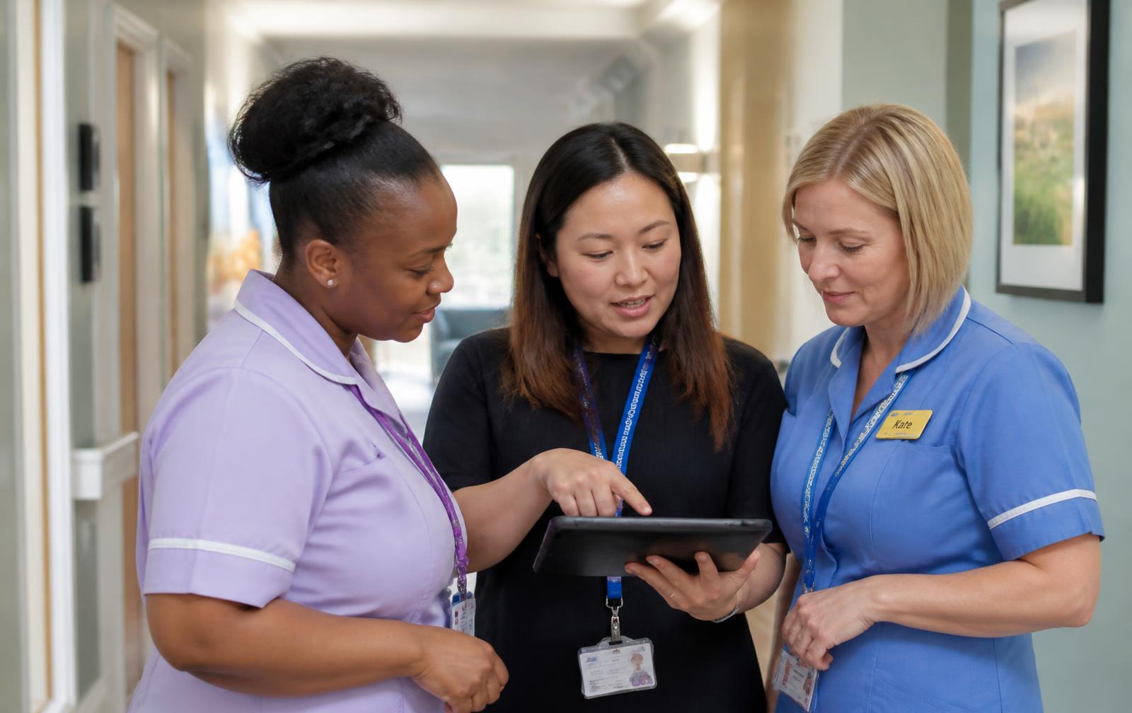 Three healthcare colleagues reviewing a handover on a tablet in a care home corridor.