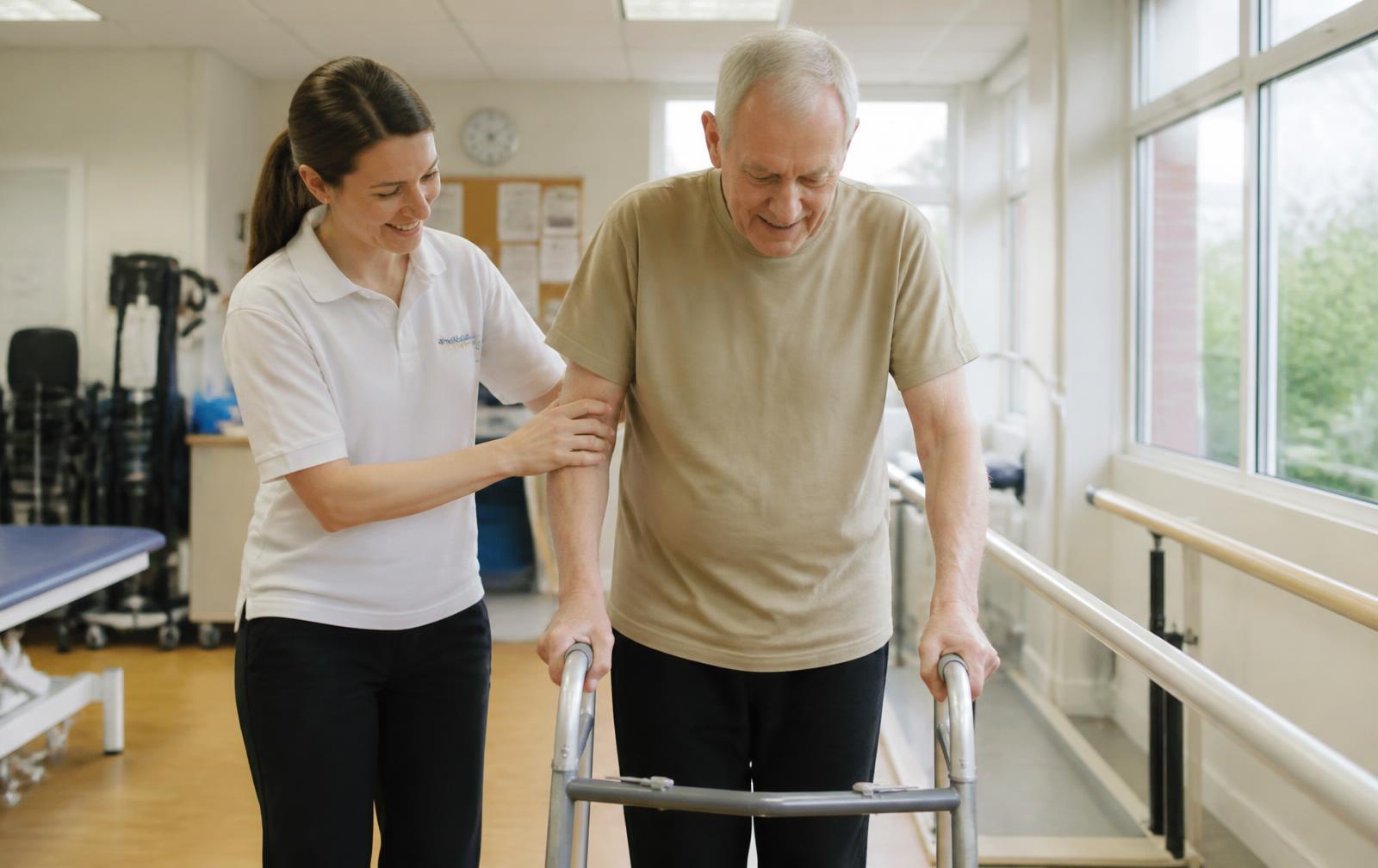 A physiotherapist supporting an older man walking with a frame in a rehabilitation room.